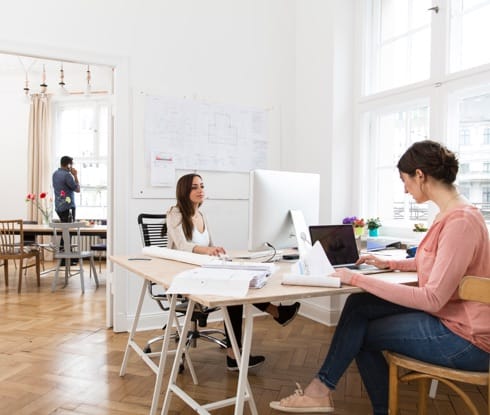 Two women work at a desk, one on a laptop computer, the other on a desktop computer.