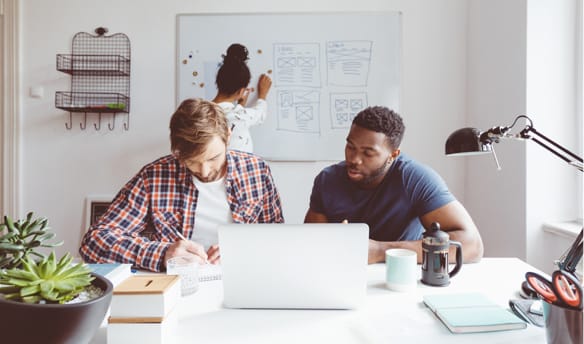 Two employees work from a laptop computer while another writes on a whiteboard.