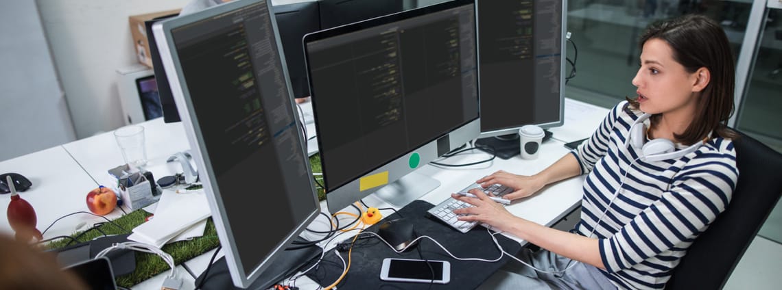 A woman types on a keyboard, looking at data spread across three computer screens. 