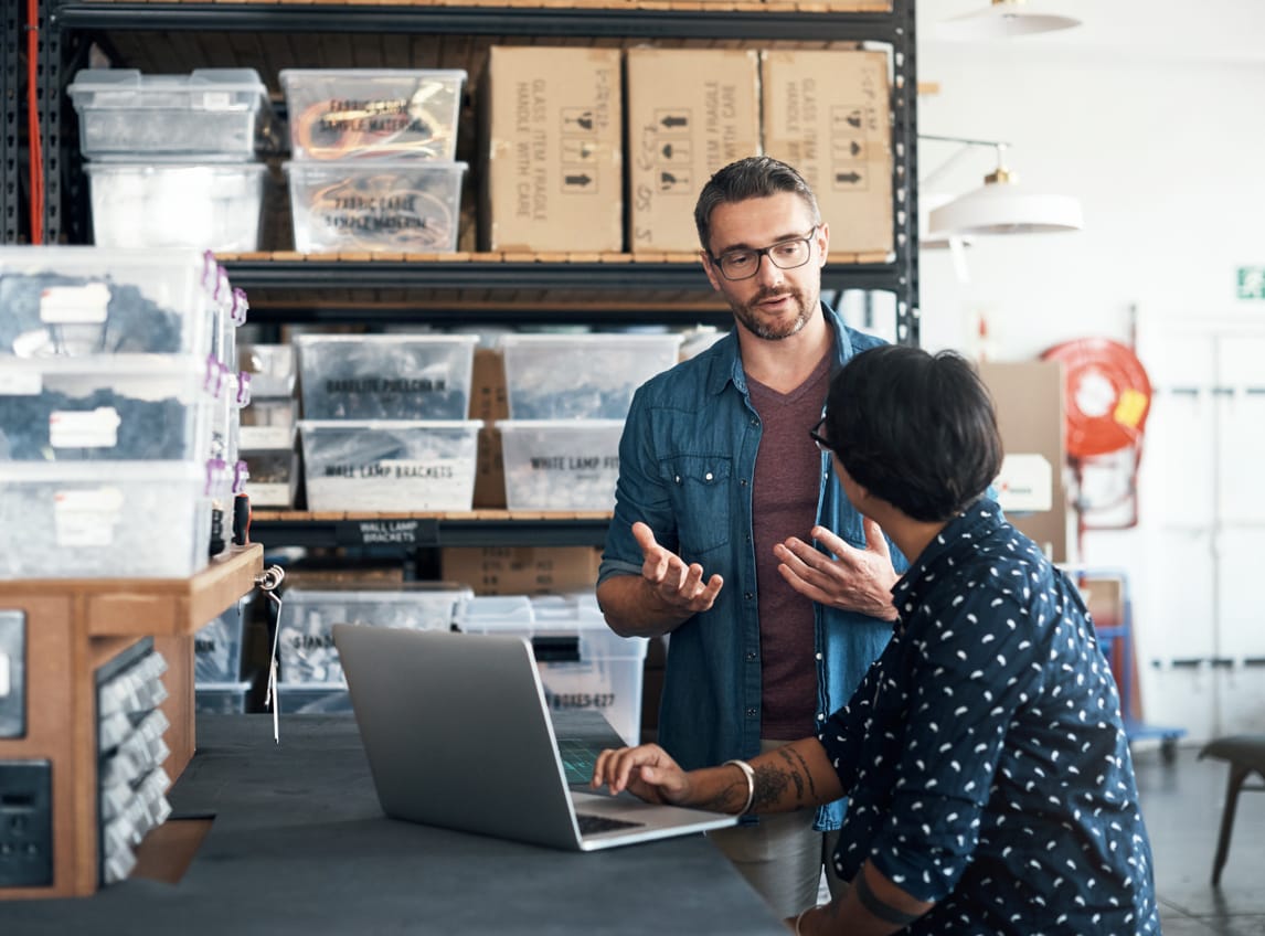 A man and woman converse in a warehouse referencing an open laptop sitting on a countertop.