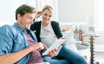 A man and a woman smile as they sit on a couch and look at a tablet screen